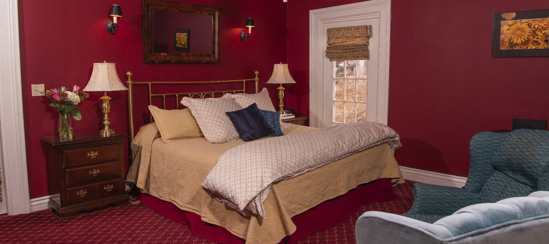 King sized brass bed in a red wallpaper and carpet in a guestroom that has a seating area with two blue chairs.